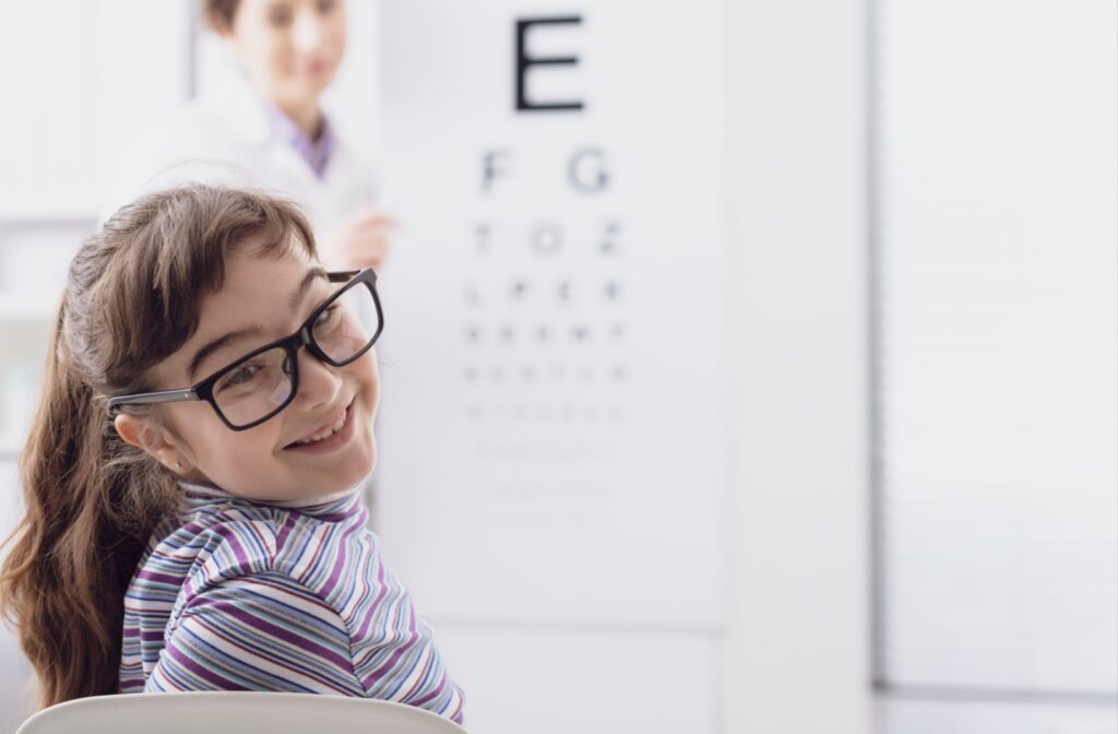 Happy young girl with glasses sitting in an optometrist's office, eye exam chart in the background.