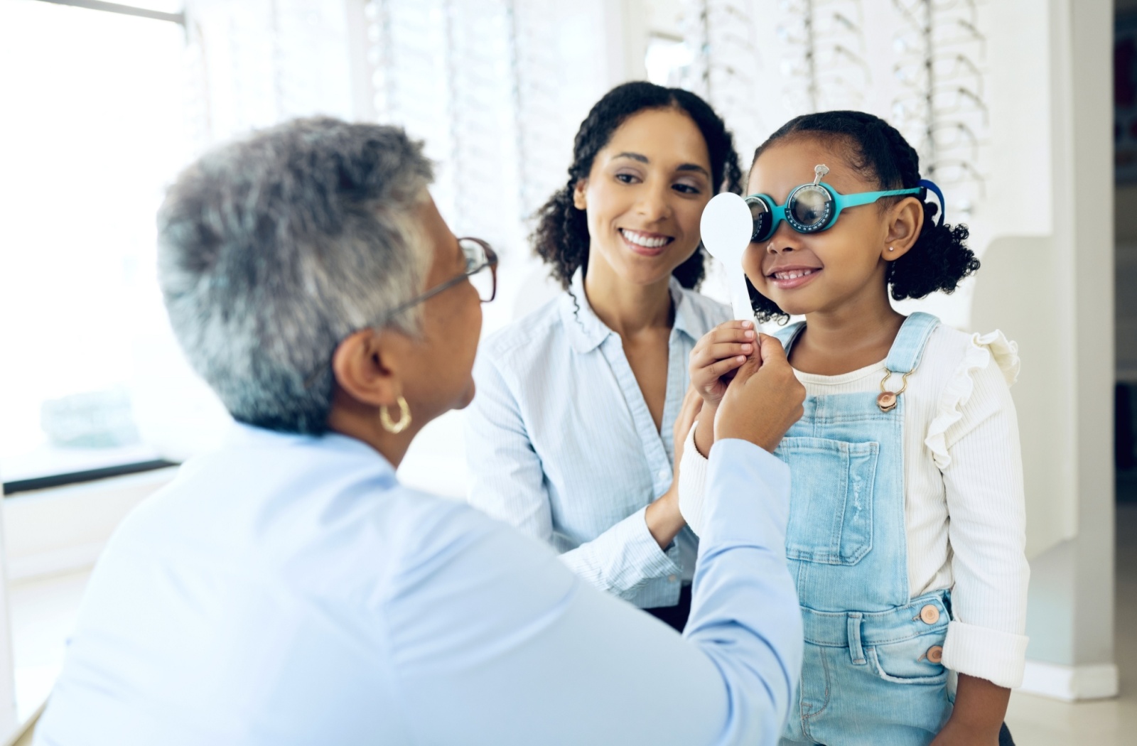 Optometrist adjusting special eyeglasses on a young girl during an eye exam, with her mother smiling beside them.