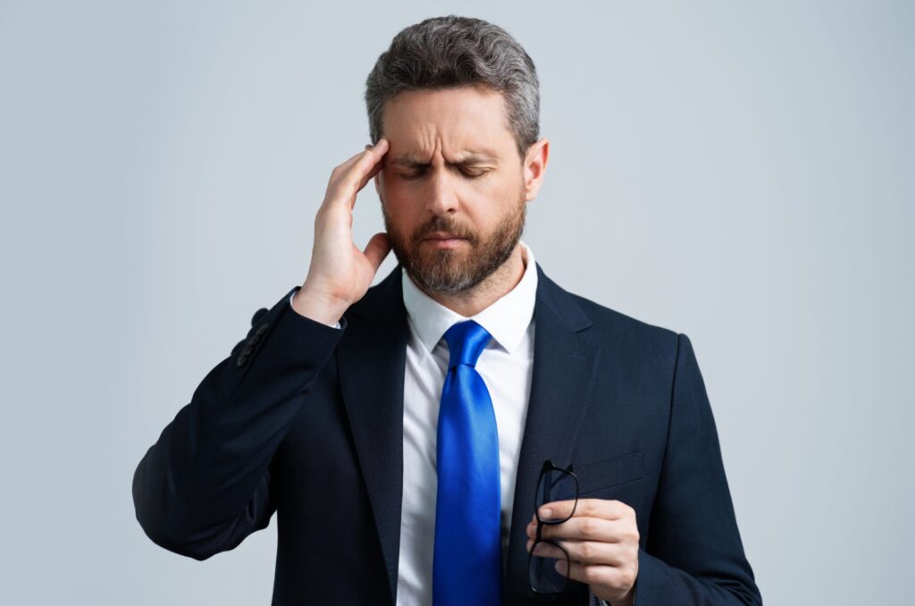 A man in a business suit and tie holding his temple with his right hand from a headache and glasses in his left hand.