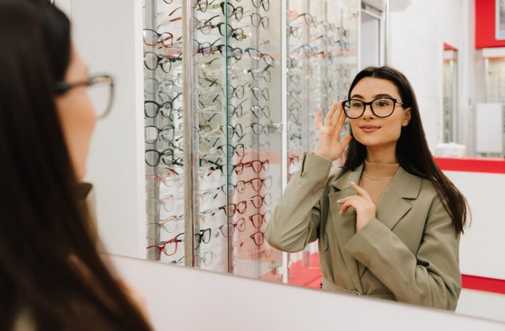 A person stands in front of a mirror and tries on glasses while a stand of trendy frames sits in the background.
