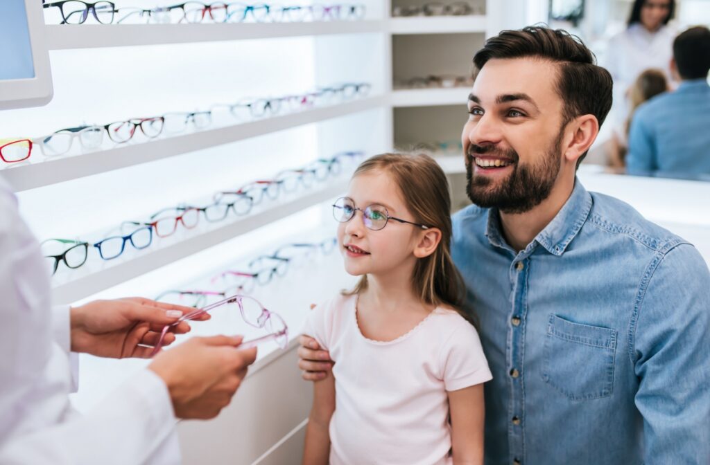 A young child and her parent stand together in an optical store, smiling as an optician shows them a pair of eyeglasses from