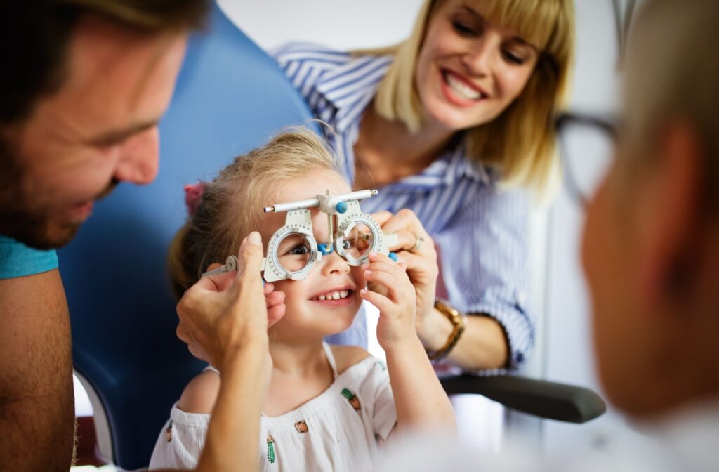 A young child smiles while sitting in an optometrist’s chair, wearing a phoropter for an eye exam, with parents helping and smiling nearby.