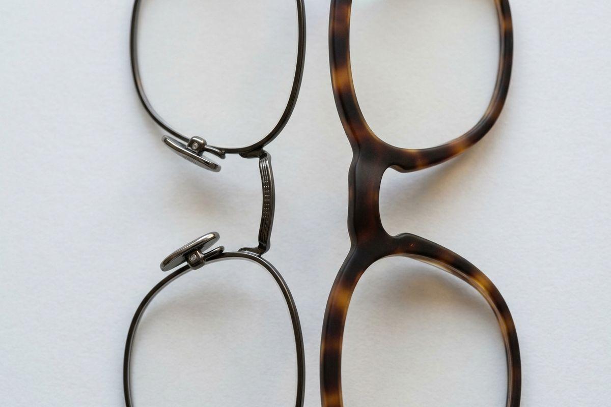 Close-up of two eyeglass frames on a white surface: a thin metal frame with nose pads beside a thick tortoiseshell plastic frame.